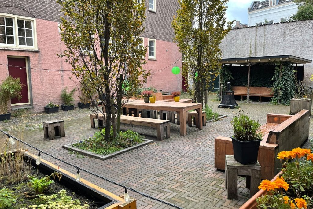 De binnentuin van Nicolaasstraat staan twee grote bomen in het midden met daartussen een grote tafel en banken. Aan de rechterkant staat een pot op een tafel met een houtenbank daarnaast. Achterin is een afdakje met een bank daar onder.